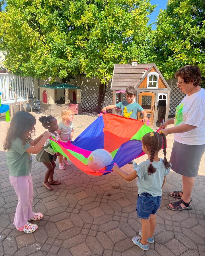 Pari leads a group of toddlers in parachute play in the sunny back yard